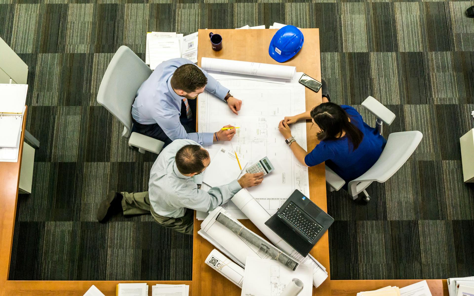 Overhead view of a team working around a table covered with laptops and plans.