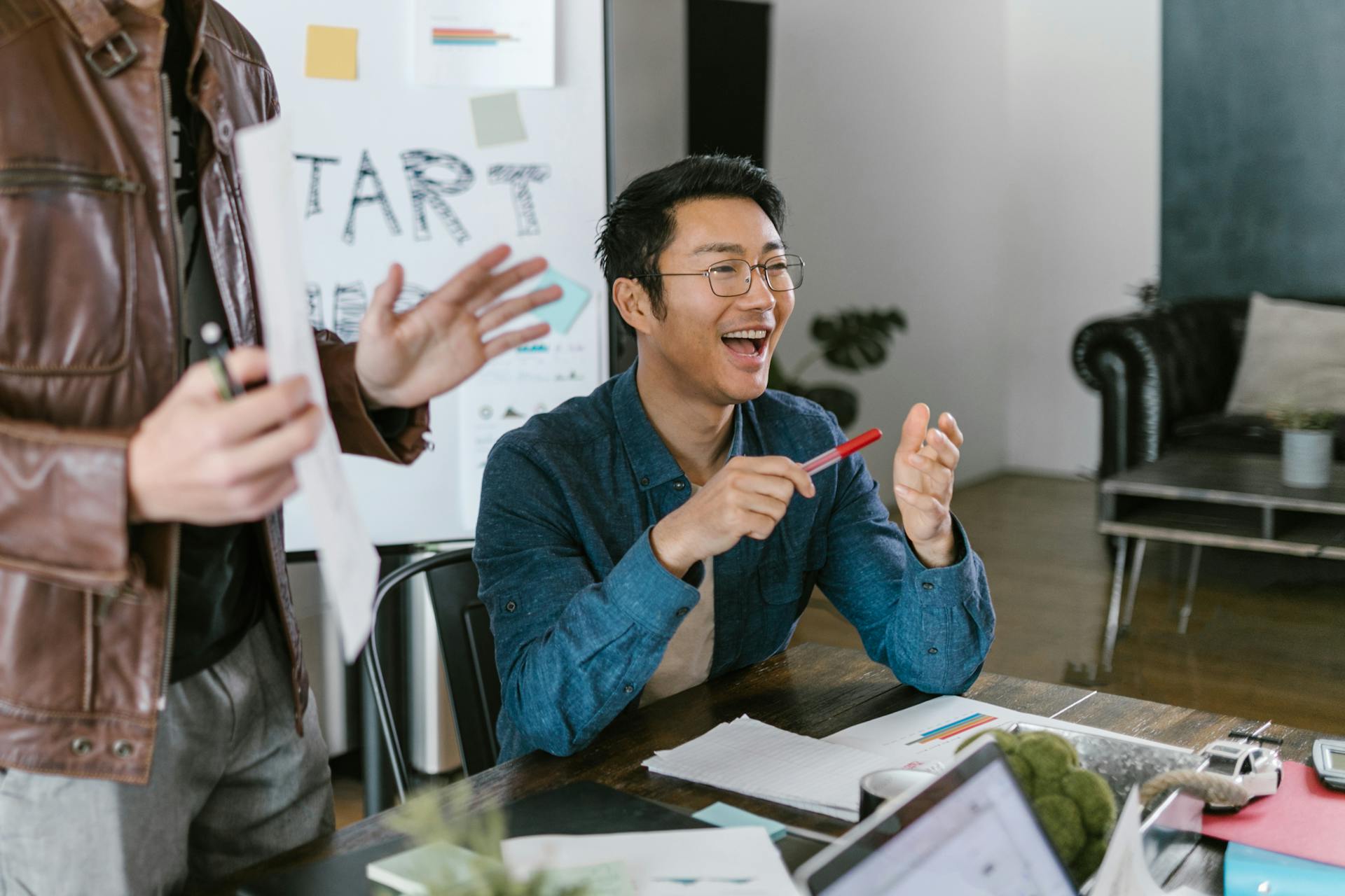 A man laughs at his desk while a colleague presents in front of a whiteboard.